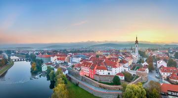 A picture of Pisek with the river and its bridges on the left side and an idyllic impression from the medium-sized town on the right. In the background small mountains are visible in what's either the beginning of sunset or end of a sunrise.