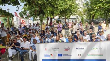 A picture of a large group of people sitting down outside listening to a presentation. In the foreground is a banner including Re-Values logo and a statement to many of the projects being funded by European funds