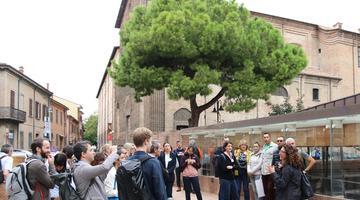Particiants on study visit A group of people standing around listening to a presenter outside. The background includes some buildings that look like heritage buildings and a beautiful tree.